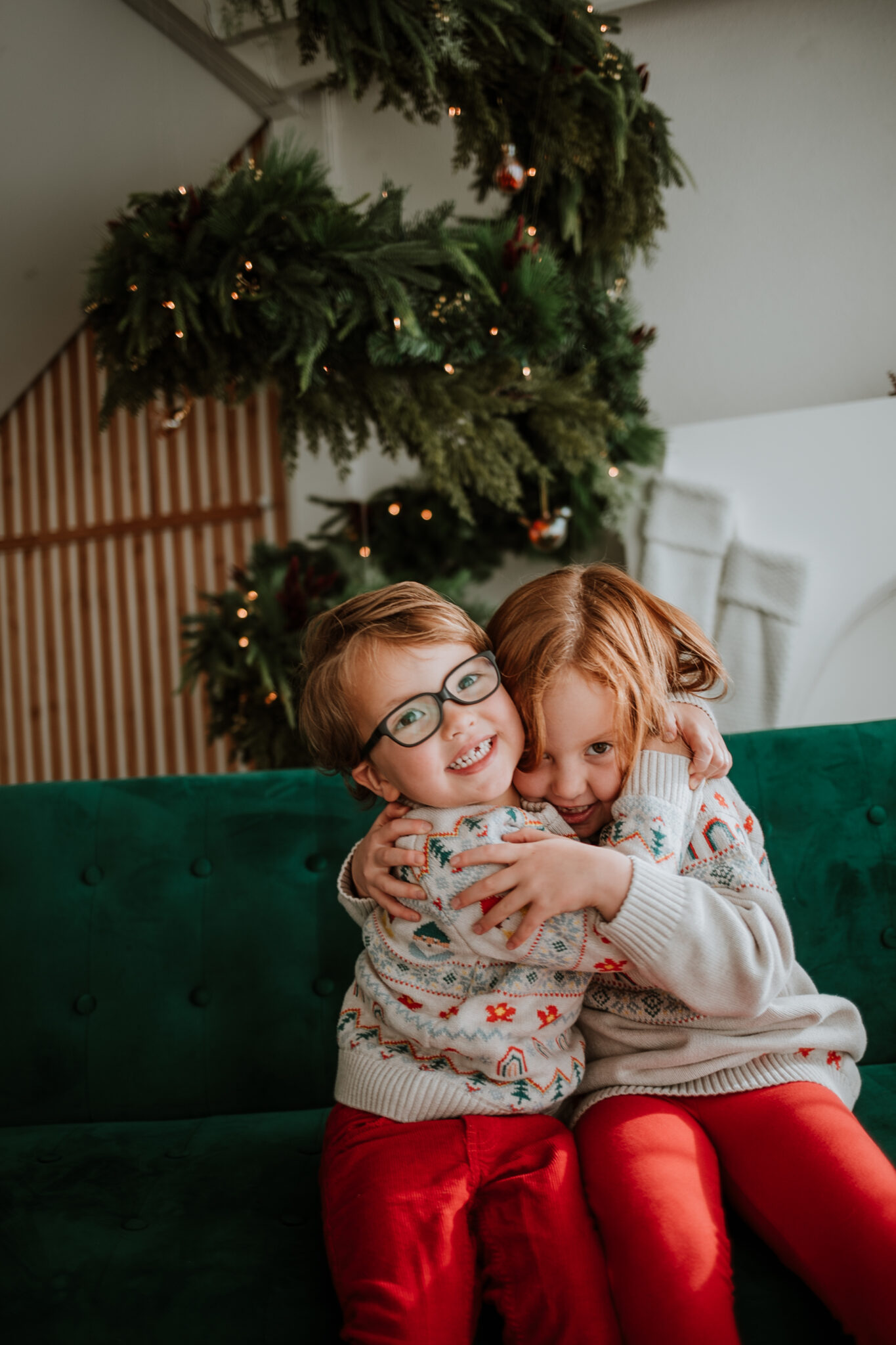Two children in cozy holiday outfits hugging and laughing in front of a glowing Christmas tree during a holiday mini session in Longmont, CO.